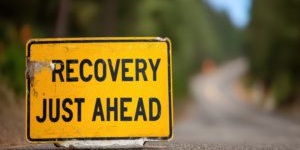 A yellow road sign with the words recovery just ahead on a blurr